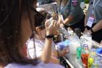 Na zdjęciu dziewczynka trzyma w ręku pipetę laboratoryjną i nabiera ciecz do probówki. Na stoliku stoją szklane butelki z kolorowymi płynami. W tle funkcjonariusze SCS.